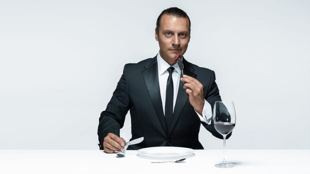 Portrait of a young man in image of maniak with knife, fork and and a piece of raw meat on a plate. Man in suit. The image in the style of Halloween