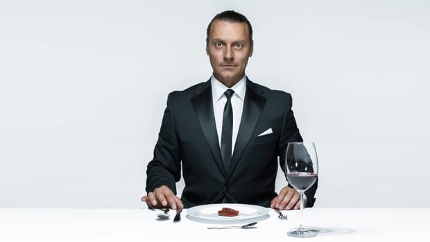 Portrait of a young man in image of maniak with knife, fork and and a piece of raw meat on a plate. Man in suit. The image in the style of Halloween