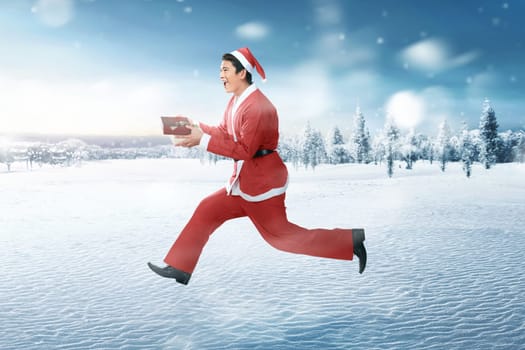 Happy asian man wearing santa claus costume with xmas gift on snowy field background