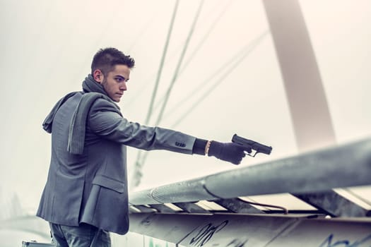 Well dressed handsome young detective or policeman or mobster standing in an urban environment aiming and firing a gun over a bridge edge