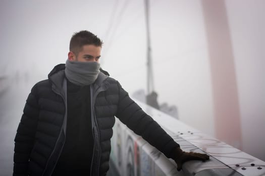 Young man in winter fashion standing outdoors in a warm overcoat and gloves with his grey scarf wound round the lower half of his face concealing his features looking away