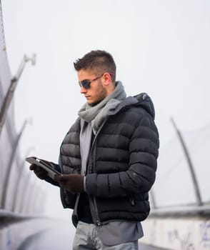 Handsome unshaven trendy man in winter fashion wearing a thick jacket, scarf and sunglasses standing looking down at a tablet computer that he is holding, side view