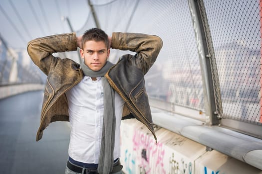 Handsome trendy young man standing on a sidewalk wearing a fashionable jacket and scarf with arms raised behind head looking at the camera