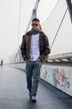 Handsome trendy young man standing on a sidewalk wearing a fashionable leather jacket and scarf in a relaxed confident pose looking at the camera