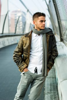 Handsome trendy young man standing on a sidewalk wearing a fashionable jacket and scarf in a relaxed confident pose looking away to a side