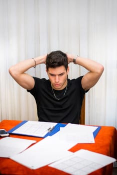 Sad or worried young man working or studying at home sitting at table with paper sheets in front of him