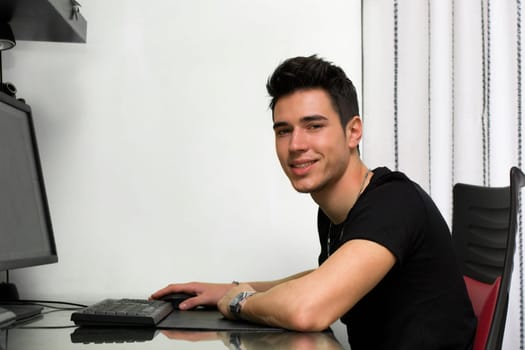 Handsome young man working or studyiing at computer at home or in office, smiling at camera