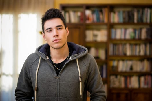 Attractive young man at home in living room with bookcase in the background, looking at camera