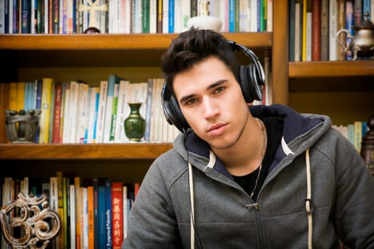 Attractive young man sitting listening to music on a set of stereo headphones in his study at home in front of a bookcase filled with books