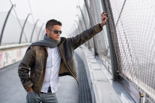 Handsome trendy young man standing on a sidewalk wearing a fashionable jacket and scarf in a relaxed confident pose looking away down the bridge