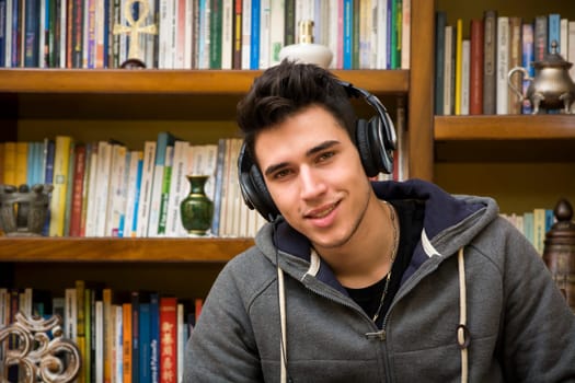 Attractive smiling young man listening to music on a set of stereo headphones in his study in front of a bookcase filled with books