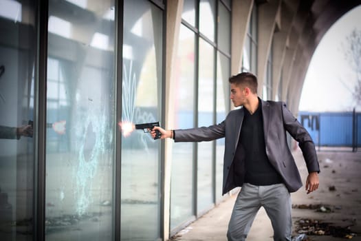 Well dressed handsome young detective or policeman or mobster standing in an urban environment aiming and firing a gun to a window glass with a determined expression, front view