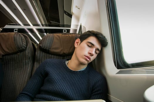 Young man sleeping while traveling on a train sitting in a passenger coach with his head resting on his hand and eyes closed