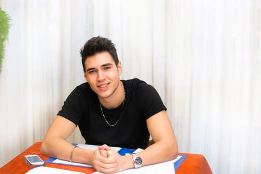 Attractive smiling young man working or studying at home sitting at table with paper sheets in front of him