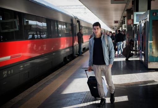 Handsome young male traveler in train station with trolley suitcase, looking at camera