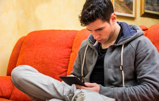 Handsome young man at home reading with ebook reader sitting on a couch
