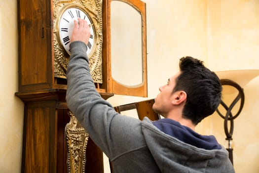 Young man checking the pendulum and winding the mechanism on a vintage long case clock standing with his back to the camera as he works