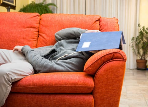 Over-worked, tired young man at home sleeping instead of working or studying, resting with head covered by book