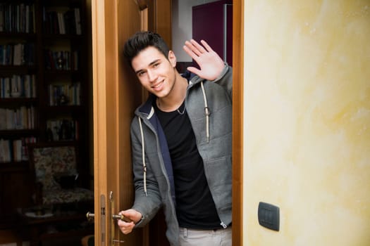 Smiling young man getting out of door waving at the camera with a friendly cheerful smile as he peers around the edge of a wooden door