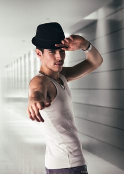 Attractive young man with black fedora hat and white tank-top in a hallway, pointing finger to the camera