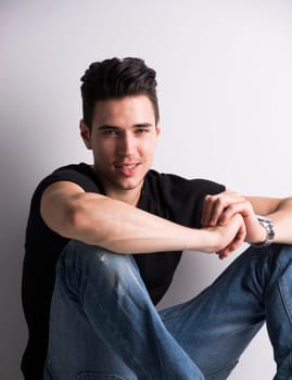 Handsome young man sitting on the floor against white wall, looking at camera smiling