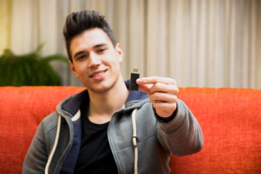 Handsome smiling young man at home showing USB key in his hand, sitting on a couch