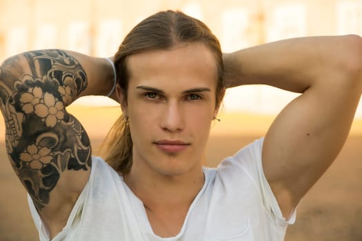 Handsome young man outdoors, with hands behind head, looking intensely at camera with confident expression