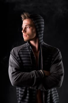 Portrait of handsome tough young man in dark hoodie on black background, looking to a side, arms crossed on chest