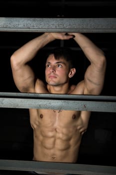 Muscular shirtless young man resting against metal structure, with arms raised above head
