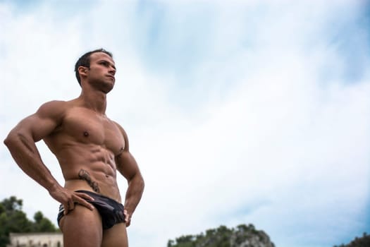 Handsome muscular young man in bathing suit shot from below, large copyspace on the sky