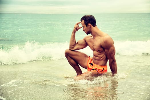 Handsome muscular young man on the beach sitting in the sea in profile pose