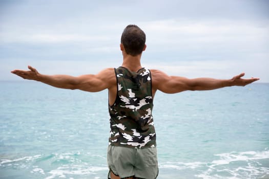 Muscular young man on the beach seen from the back, with arms open enjoying the sensation