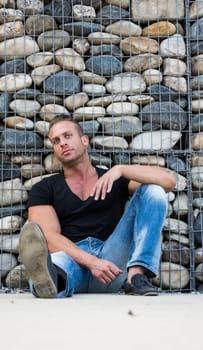Handsome muscular blond man sitting against stones wall, looking at camera