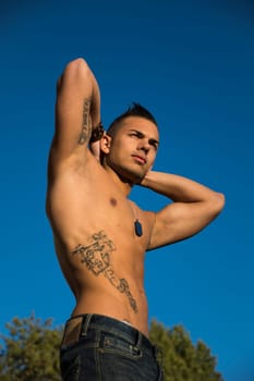 Handsome fit athletic shirtless young man against blue sky shot from below, with arms raised behind head
