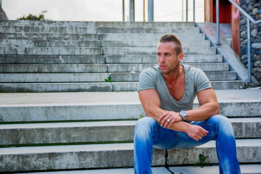 Handsome muscular blond man sitting on stair steps in city setting looking away