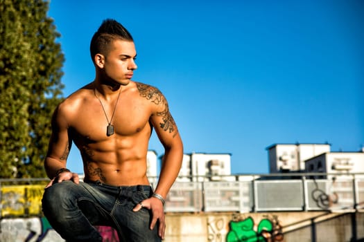 Handsome fit athletic shirtless young man in city setting kneeling and looking to a side, large copyspace
