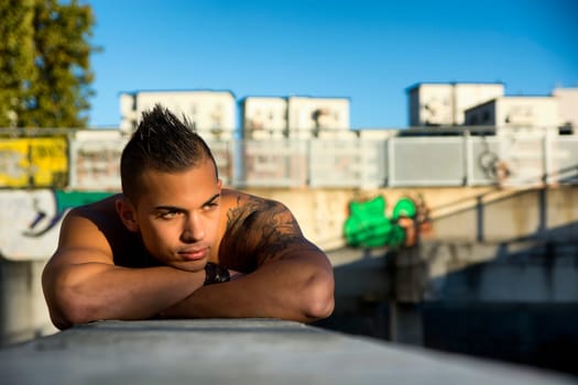 Handsome fit athletic shirtless young man in city setting leaning on concrete, looking away, large copyspace
