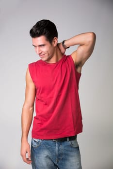 Smiling Young Man Posing in Casual Fuchsia Sleeveless Shirt and Faded Jeans with One Hand on the Nape While Glancing at the Camera. Isolated on Gray Background.