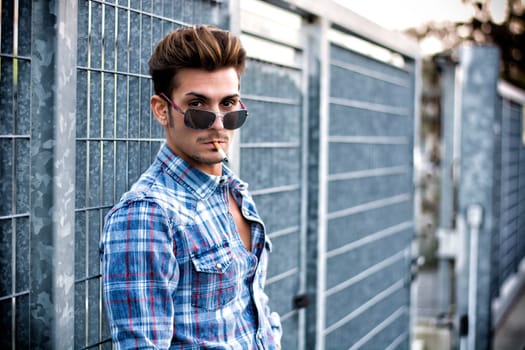 Trendy cool young man standing outside smoking, leaning against metal gate, wearing shirt and jeans