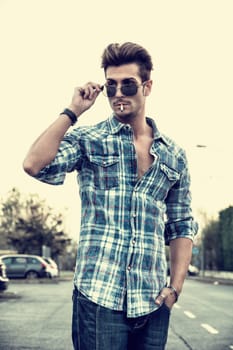 Trendy cool young man standing outside smoking, lowering sunglasses, wearing shirt and jeans