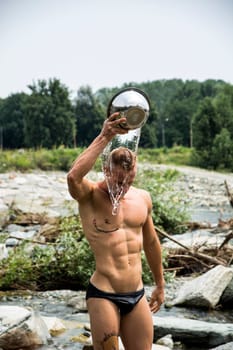 Shirtless muscular young pouring water on his head with metal bowl outdoor near river
