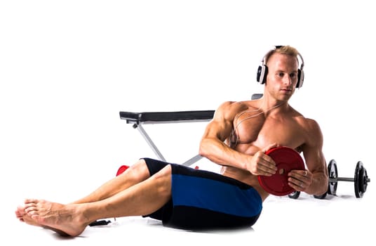 Muscular shirtless young man exercising with weights, bench, dumbbells, listening to music, isolated on white