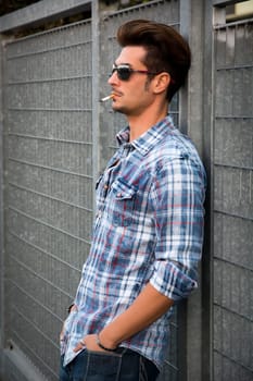 Trendy cool young man standing outside smoking, leaning against metal gate, wearing shirt and jeans