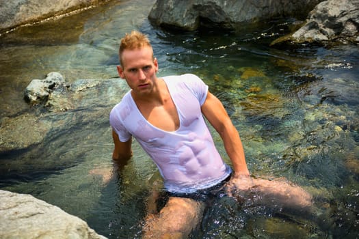 Handsome young muscle man sitting in water pond, wearing wet t-shirt looking at camera