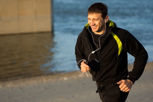 Smiling man is running with earphones outdoors on the beach.