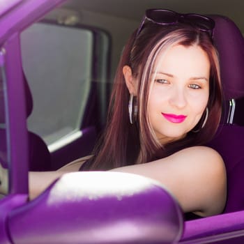 Fashionable attractive woman drives purple car and looks at herself in the mirror.