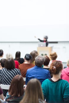 Business and entrepreneurship symposium. Female speaker giving a talk at business meeting. Audience in conference hall. Rear view of unrecognized participant in audience. Copy space on whitescreen.