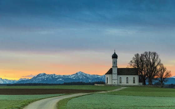 Sunrise over old chapel in green field with Alps mountains on background