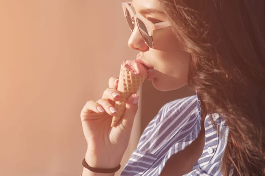 Young, beautiful brunette woman eating pink ice cream in a waffle horn. She wears glasses with dark glasses, and a shirt in a blue and white strip. Close-up photo