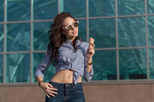 Young, beautiful brunette woman eating a pink ice cream in a waffle horn. She's in a good mood She wears glasses with dark glasses, a shirt in a white and blue strip, and dark blue jeans.